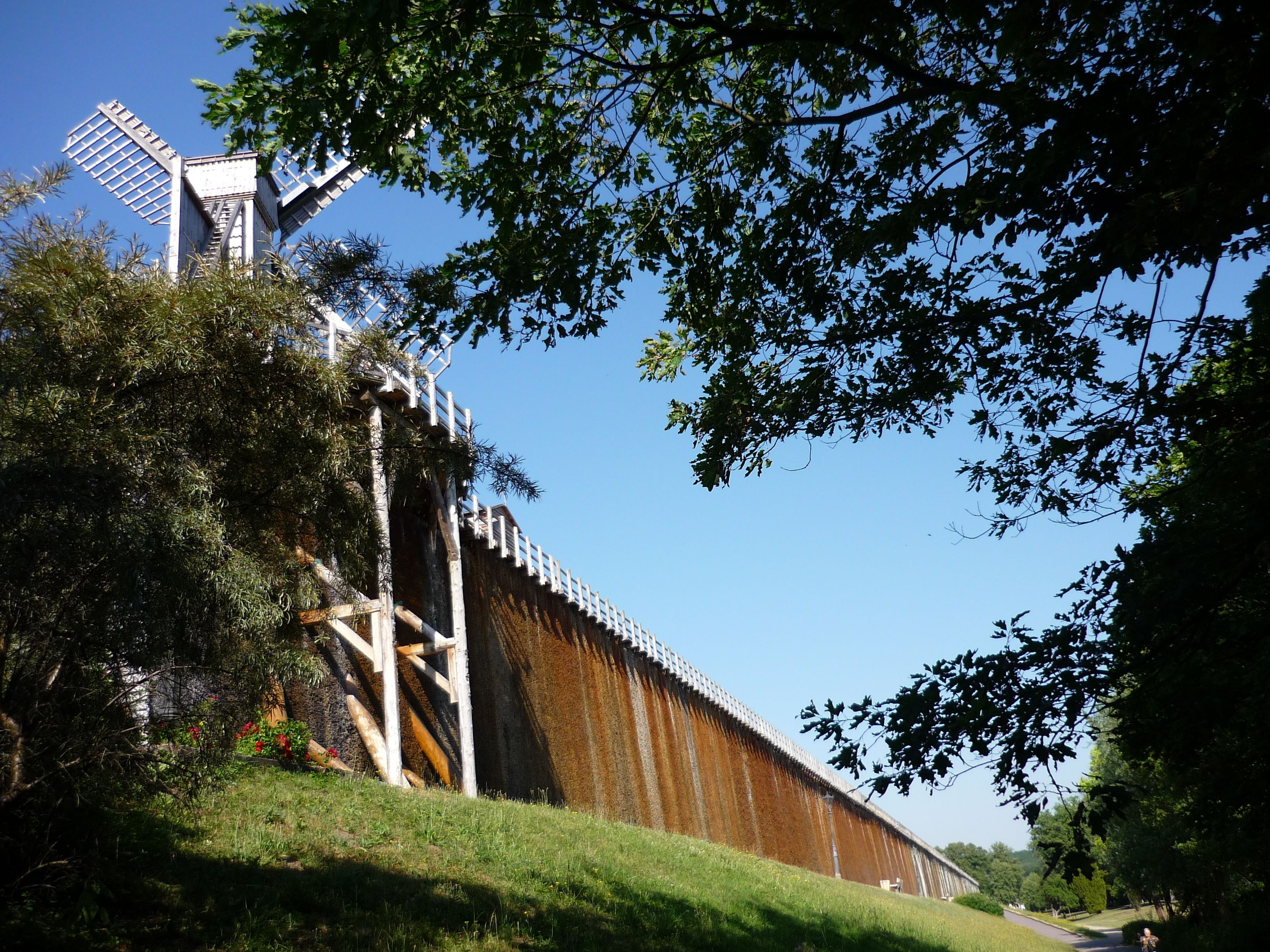 A windmill with a white structure and a rusted metal wall, surrounded by greenery and trees.