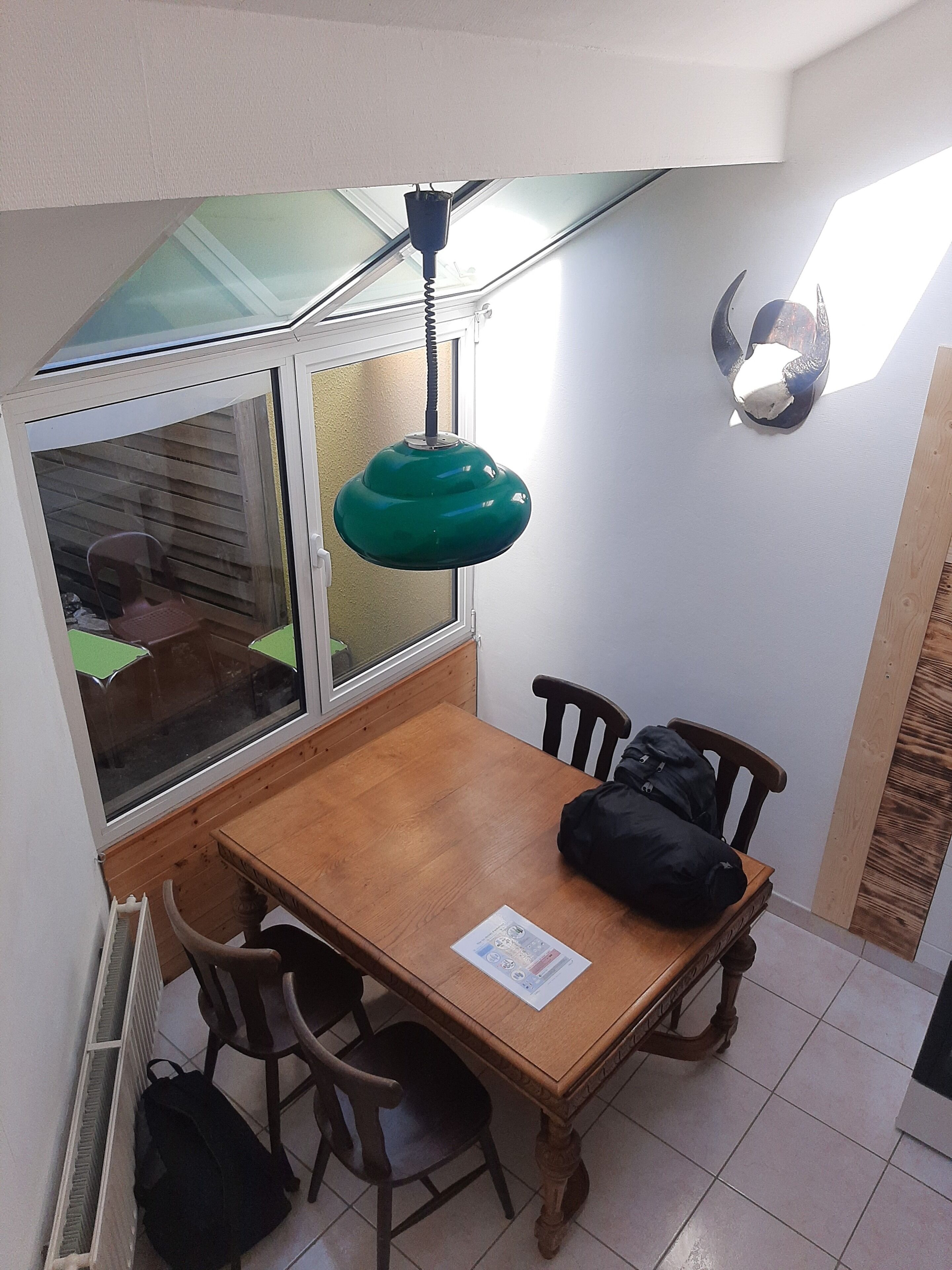 A small dining area with a wooden table, four chairs, a green pendant light, and a window with blinds.