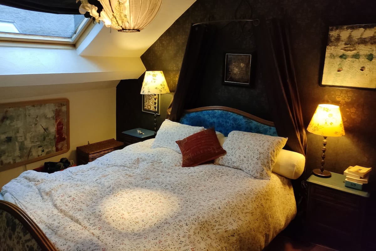 A bedroom with a large bed, two bedside lamps, a window, and a framed picture on the wall.