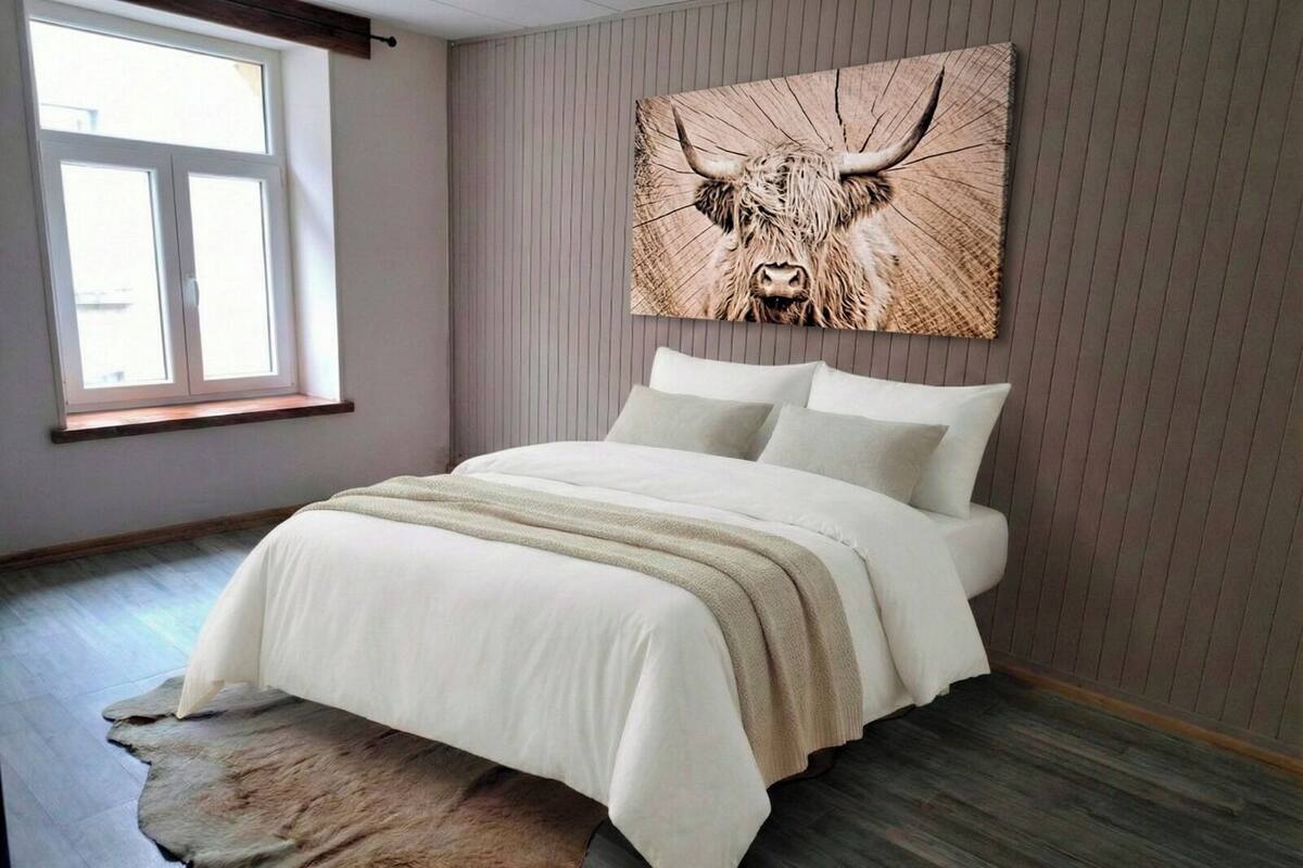 A bedroom with a large bed, a fur rug, a wooden floor, a window, and a wall-mounted artwork of a bull.