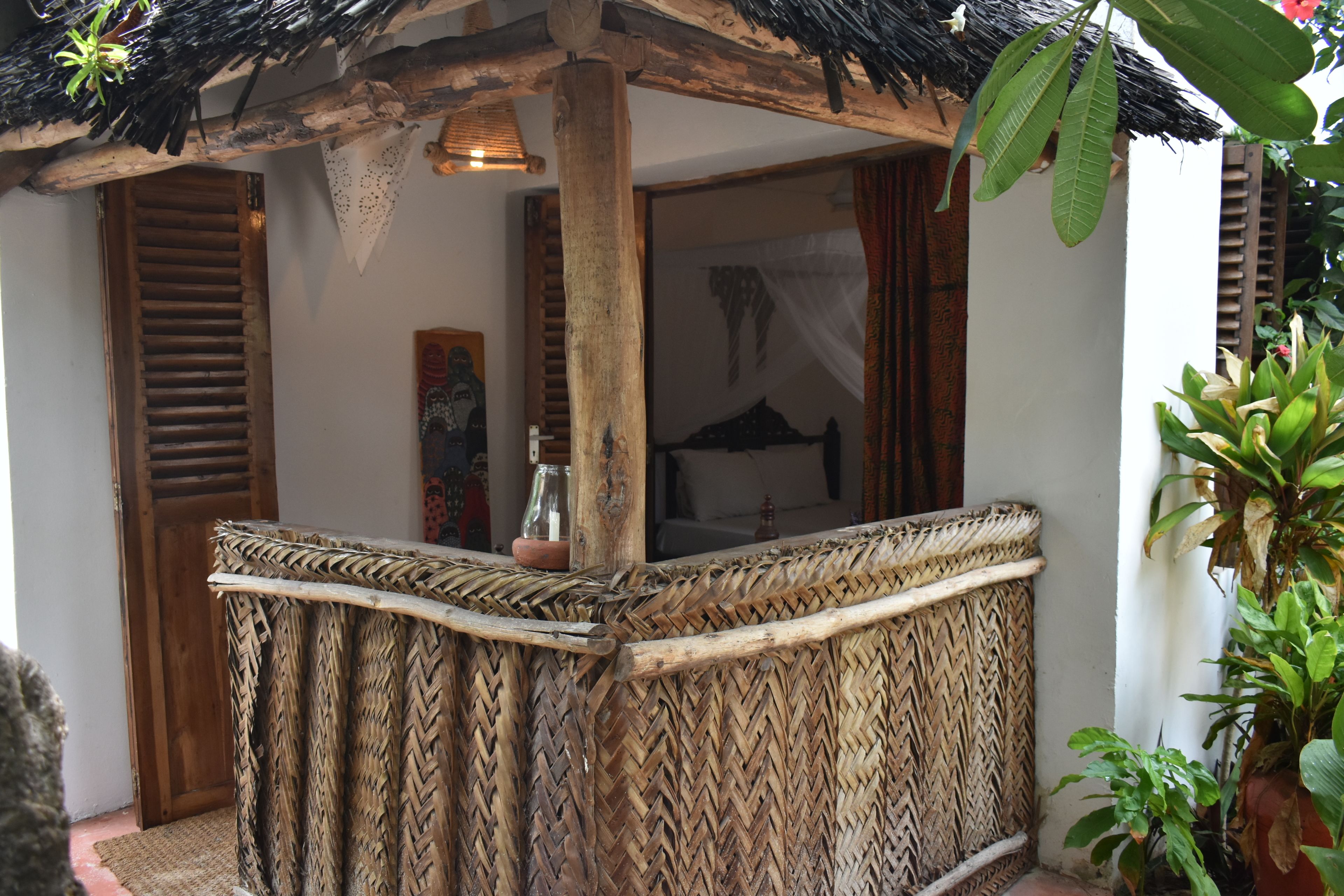A thatched-roof outdoor seating area with a woven bench, wooden door, and a view of the interior.