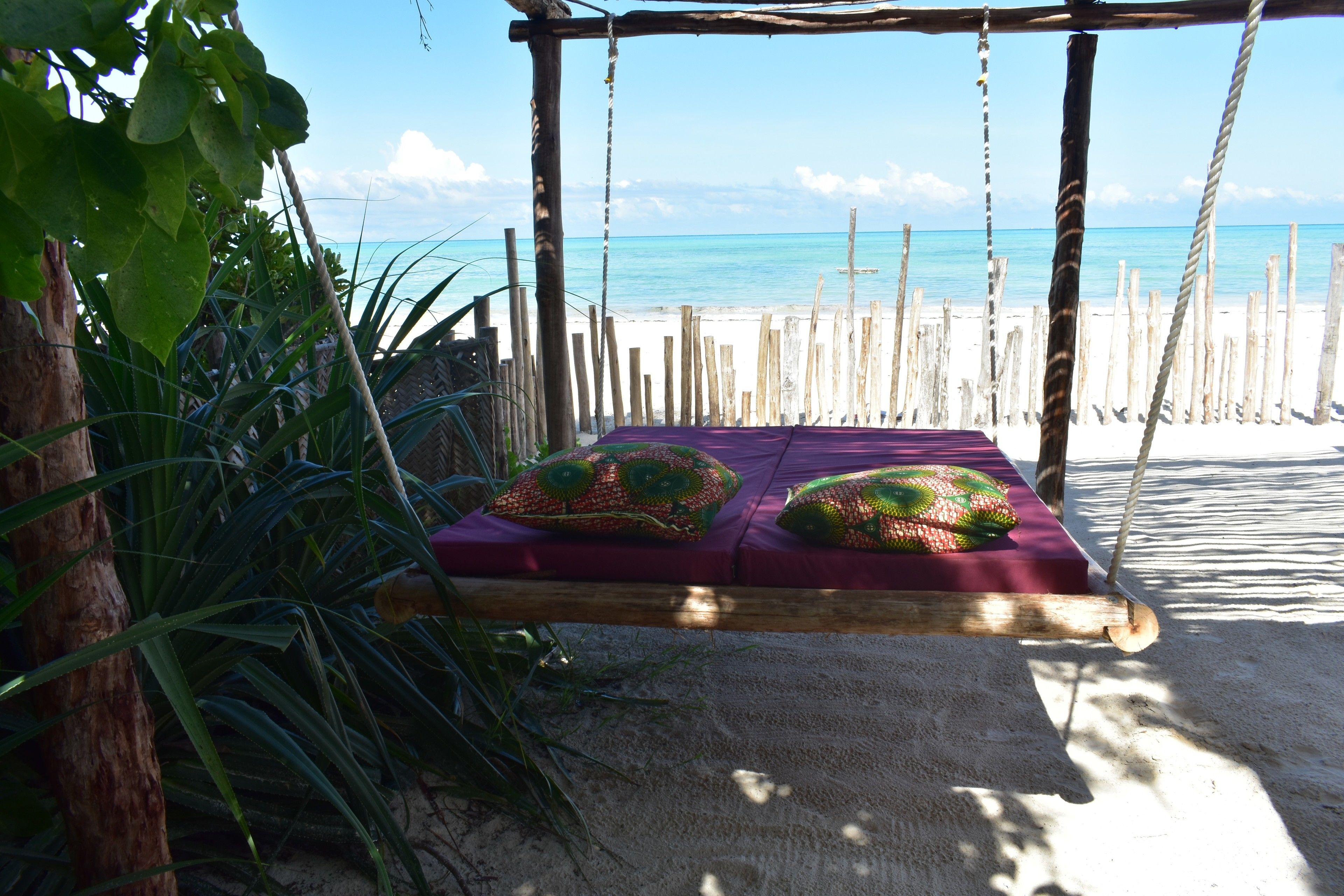 A wooden swing bed with a purple mattress and green pillows, overlooking a beach with a wooden fence and clear blue water.