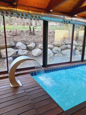 A swimming pool with a waterfall feature, surrounded by a wooden deck and large windows offering a view of a landscaped garden with rocks and trees.