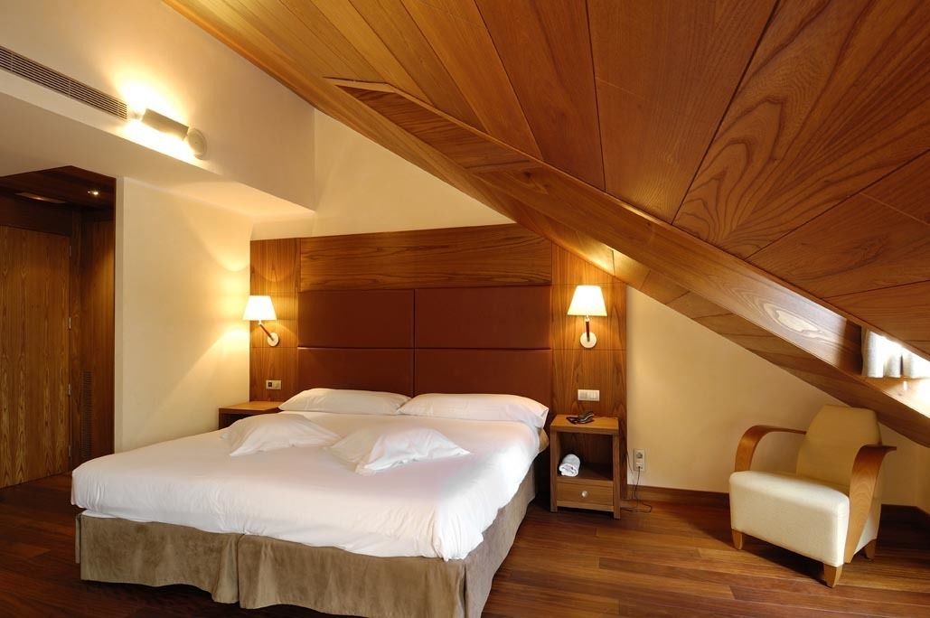 A bedroom with a wooden ceiling, a bed with white linens, two bedside lamps, a nightstand, and a chair.