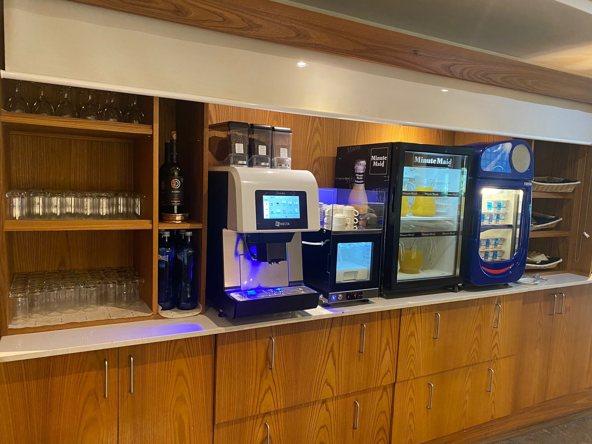 Una estación de bebidas autoservicio con varios dispensadores, incluyendo café y té, en un ambiente moderno con armarios de madera.