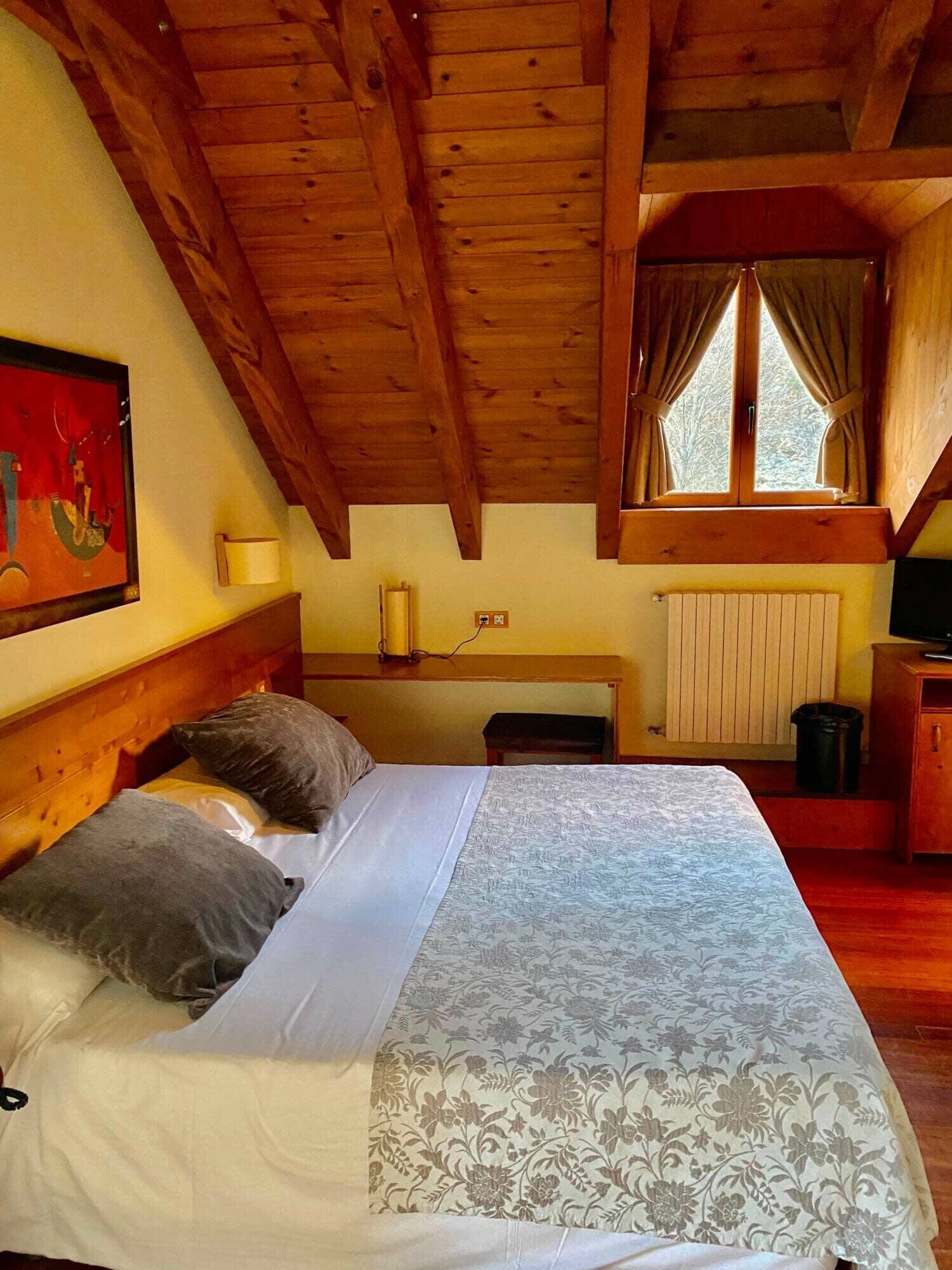 A wooden cabin bedroom with a bed, pillows, a desk, a lamp, and a window with curtains.
