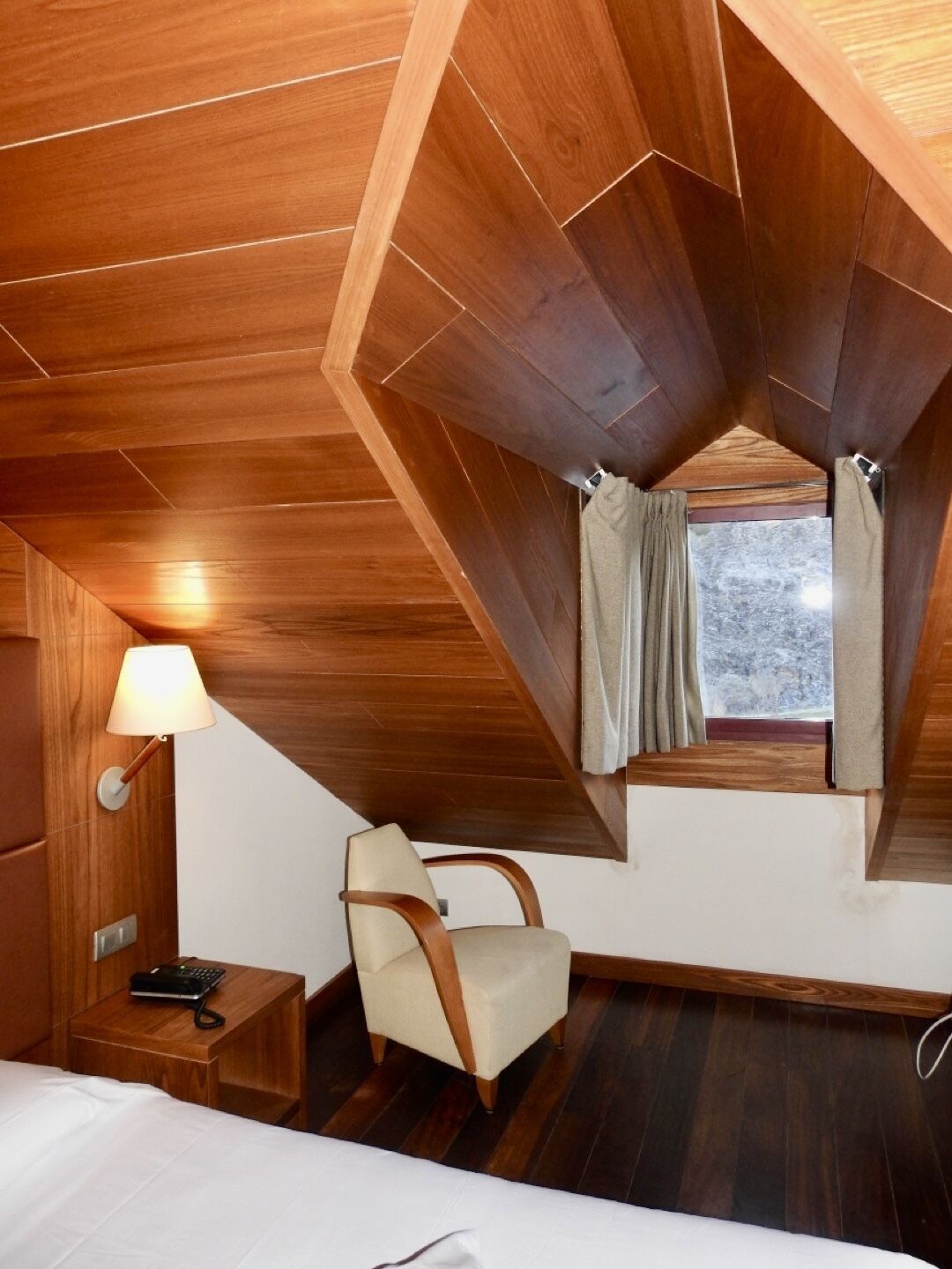 Une pièce avec un plafond en pente, un lit, une chaise, une table de chevet avec une lampe et une fenêtre avec des rideaux.