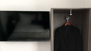 Habitación de hotel con televisor de pantalla plana en la pared, armario de madera con perchero, una mesita con tetera, tazas y bandeja, y un otoman gris.