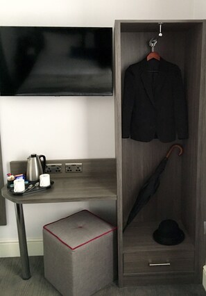 A hotel room with a flat-screen TV mounted on the wall, a wooden wardrobe with a hanging coat, a small table with a kettle, cups, and a tray, and a grey ottoman. - The Beverley Hotel (London)