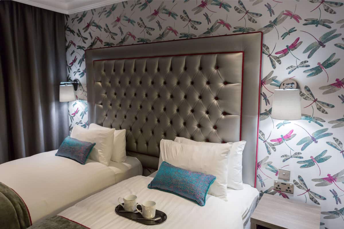 A hotel room with a tufted headboard, two beds with white linens, decorative pillows, a tray with two mugs, and a wall with a dragonfly pattern.