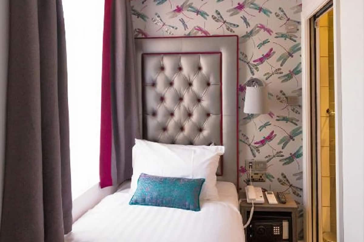 A hotel room with a tufted headboard, a bed with white linens and a teal pillow, a nightstand with a lamp, and a wall with a floral wallpaper pattern.