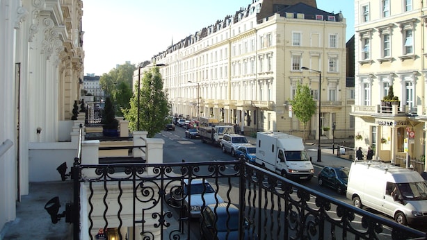 Vista de la calle con una hilera de edificios europeos clásicos, varios vehículos estacionados y cielo despejado.