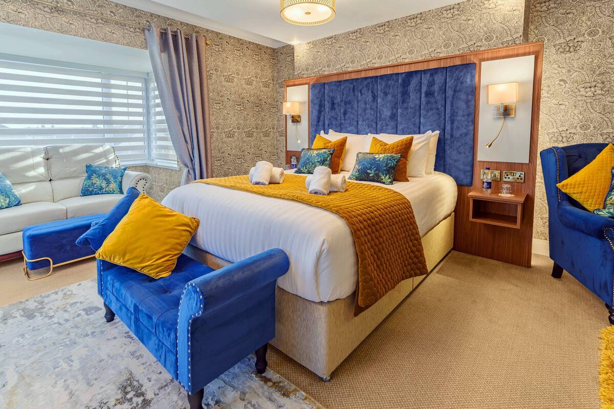 A hotel room with a large bed, two blue armchairs, a sofa, and a patterned carpet.