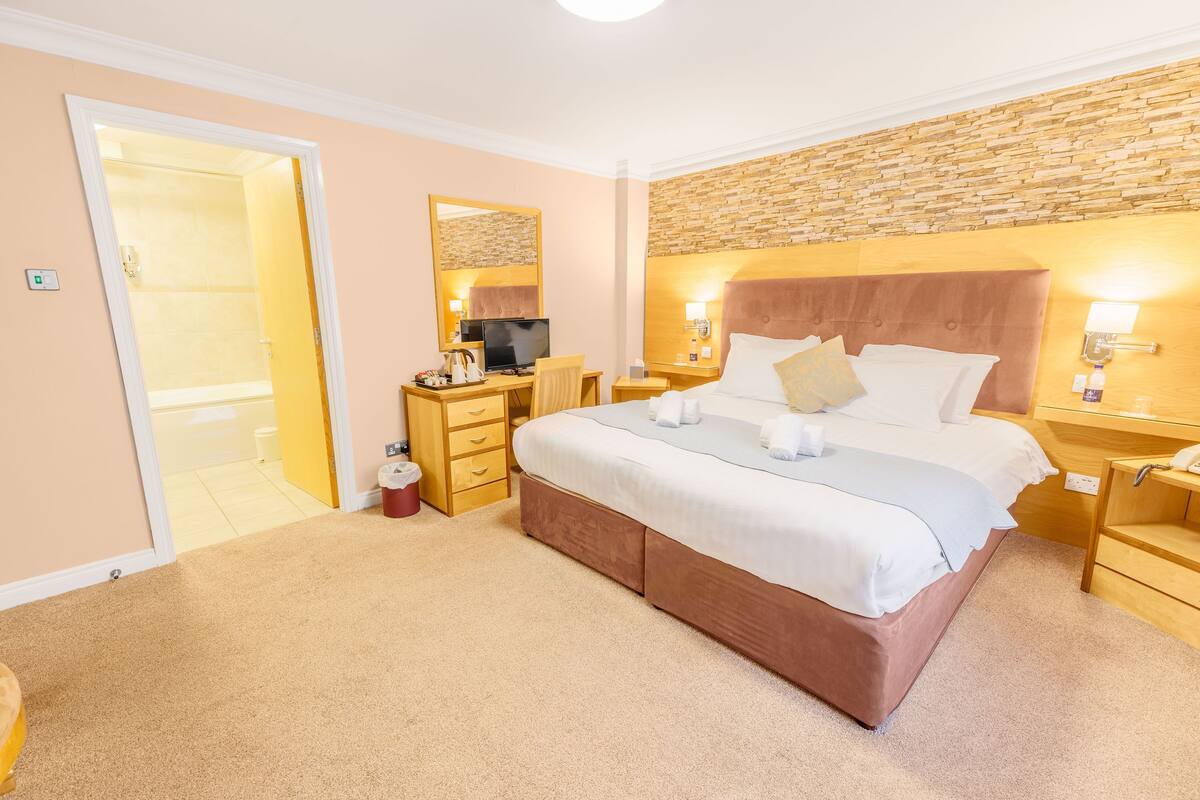 A hotel room with a large bed, a desk with a computer, a chair, and a bathroom visible through an open door.