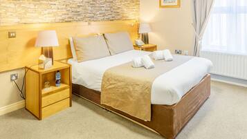 Quarto de hotel com parede de tijolos, cabeceira de madeira, cama com lençóis brancos e manta bege, duas mesas de cabeceira com luminárias, um criado-mudo com abajur, janela com cortinas e radiador.