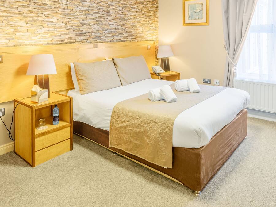 Quarto de hotel com parede de tijolos, cabeceira de madeira, cama com lençóis brancos e manta bege, duas mesas de cabeceira com luminárias, um criado-mudo com abajur, janela com cortinas e radiador.