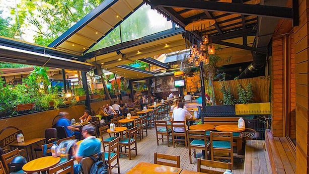 Un café en plein air avec des tables et des chaises en bois, entouré de verdure et doté d’un espace couvert agrémenté de guirlandes lumineuses.