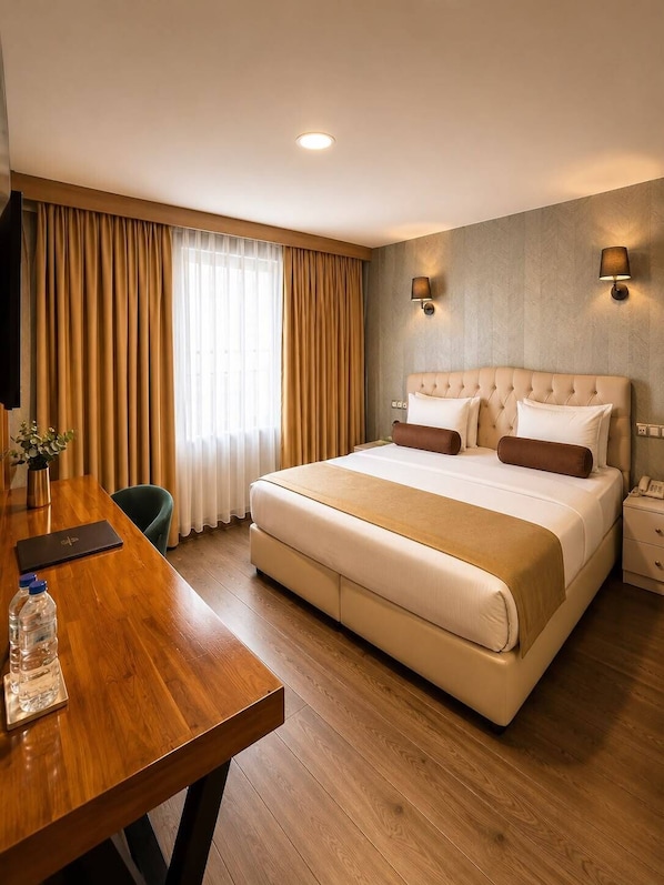 Habitación de hotel con una cama grande, un escritorio de madera, una silla, una mesita de noche con lámpara y una ventana con cortinas.