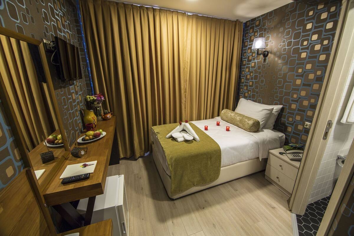 A hotel room with a bed, bedside table, and a desk with a chair. There is a window with curtains, a wall with a geometric pattern, and a lamp on the wall.