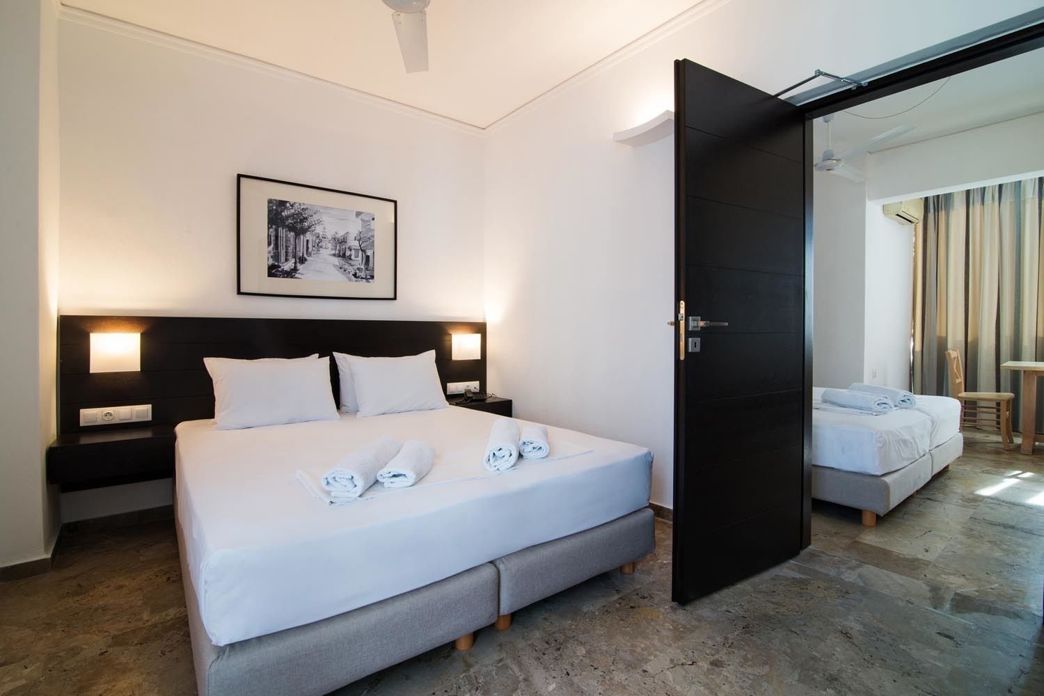 A hotel room with a large bed, a bedside table, a nightstand, a framed picture on the wall, and a door leading to another room.