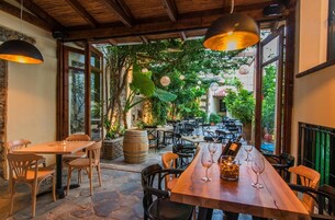 An outdoor dining area with stone flooring, wooden tables and chairs, and hanging pendant lights.