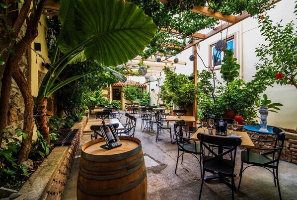 Un café al aire libre con mesas y sillas, rodeado de abundante vegetación y un rústico muro de piedra.