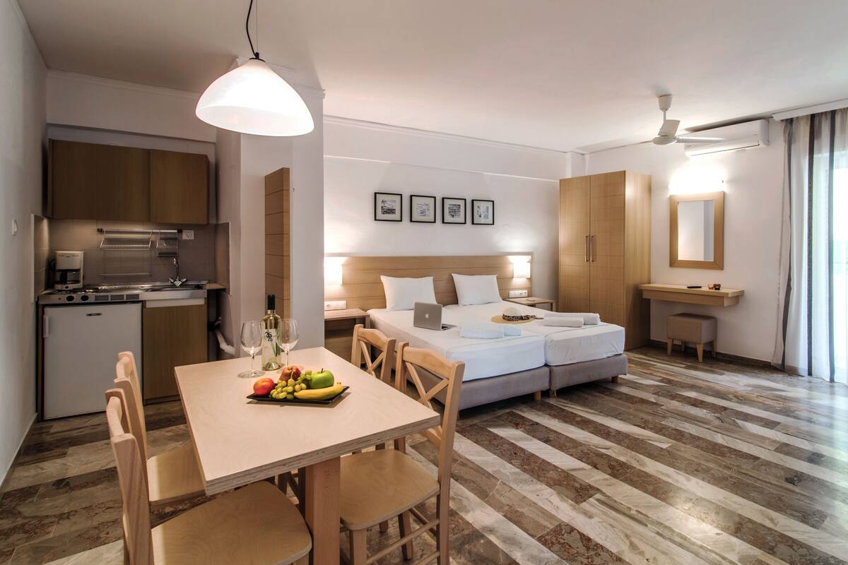 Una habitación de hotel moderna con cocina, una mesa de comedor con fruta, una cama con un ordenador portátil y un baño con espejo.