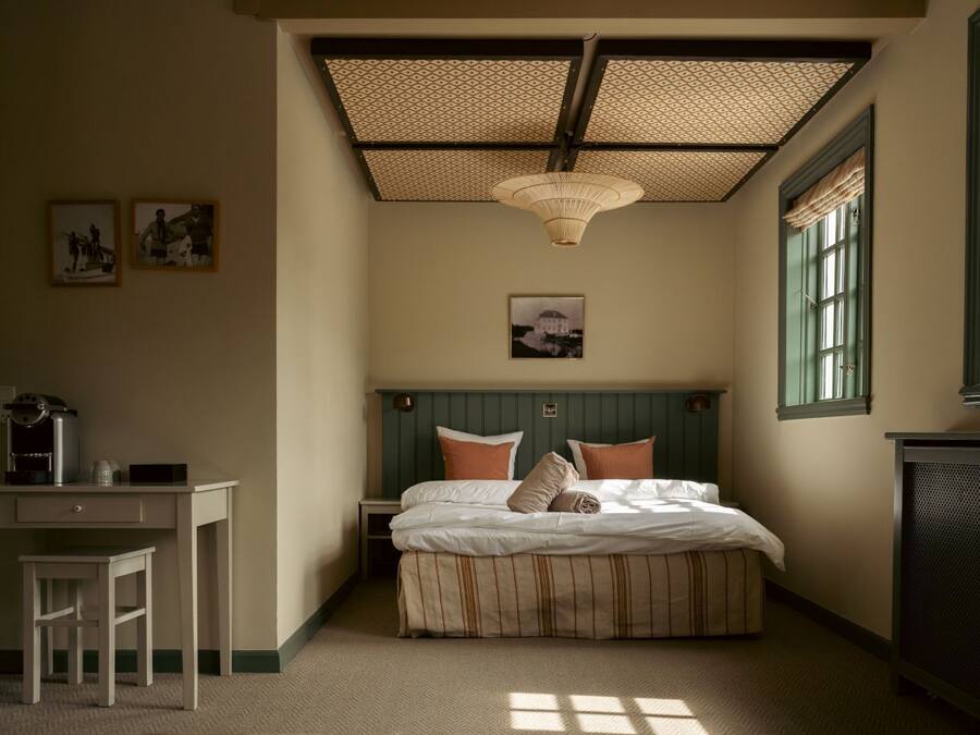 Un dormitorio con cama, escritorio y silla. Hay dos ventanas con cortinas, un ventilador de techo y una lámpara fijada a la pared.