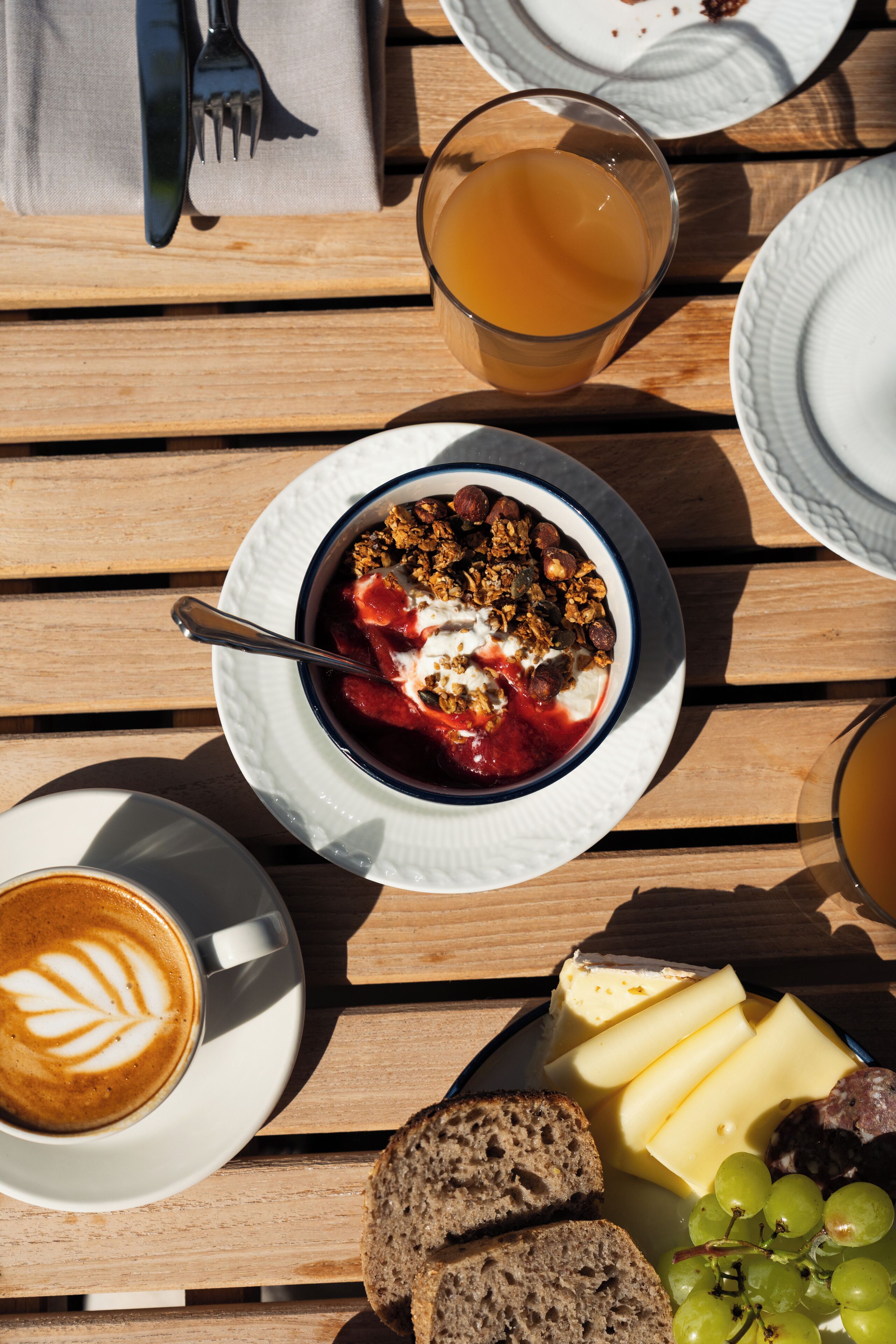 Ein Frühstücksbuffet mit Joghurt, Granola und Beeren, einem Glas Orangensaft, Käse und Trauben, Brotscheiben und einer Kaffeetasse mit Latte Art.