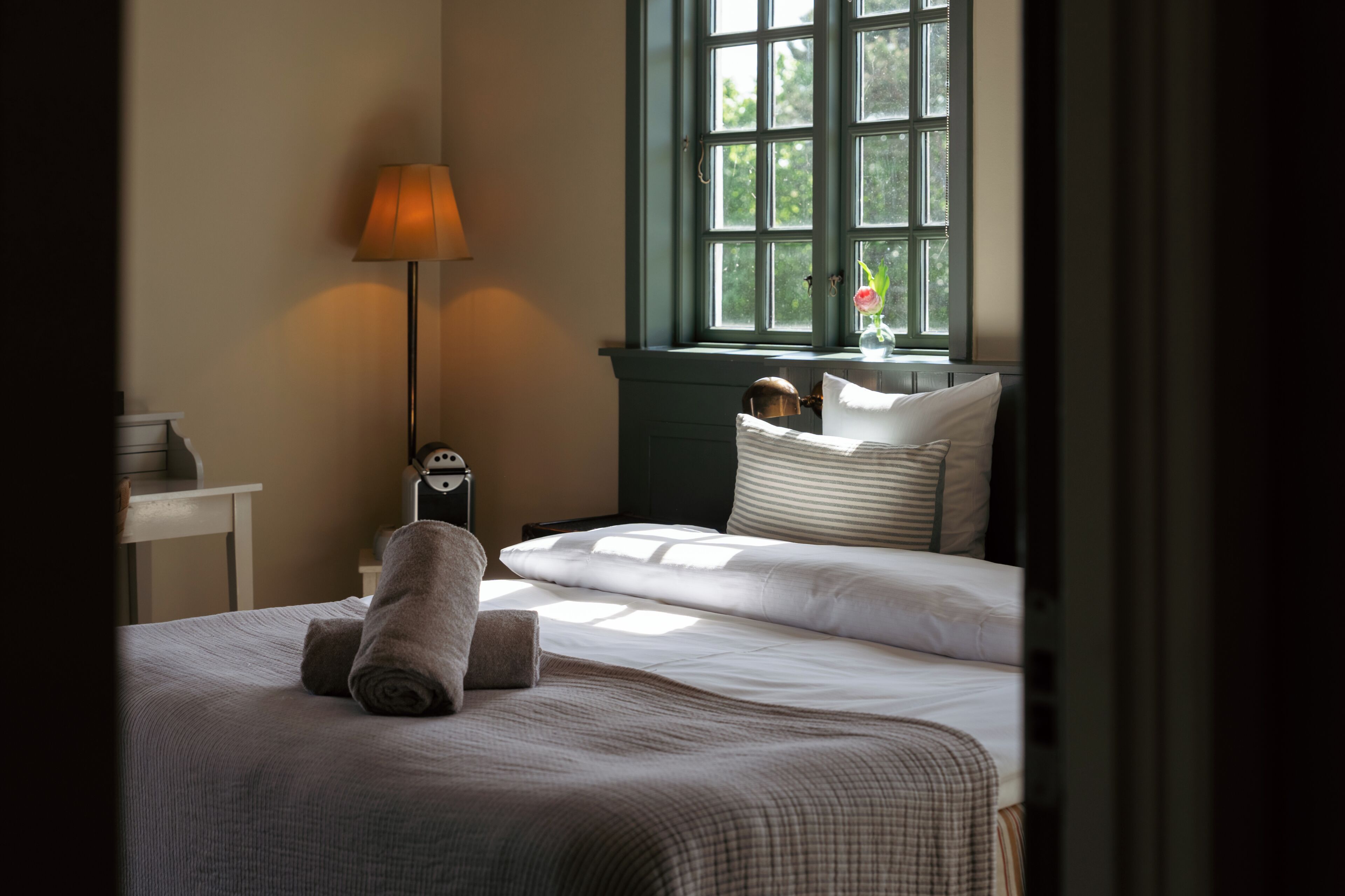 Una cama bien hecha con una almohada, una lámpara de noche y una ventana con vistas a la vegetación.