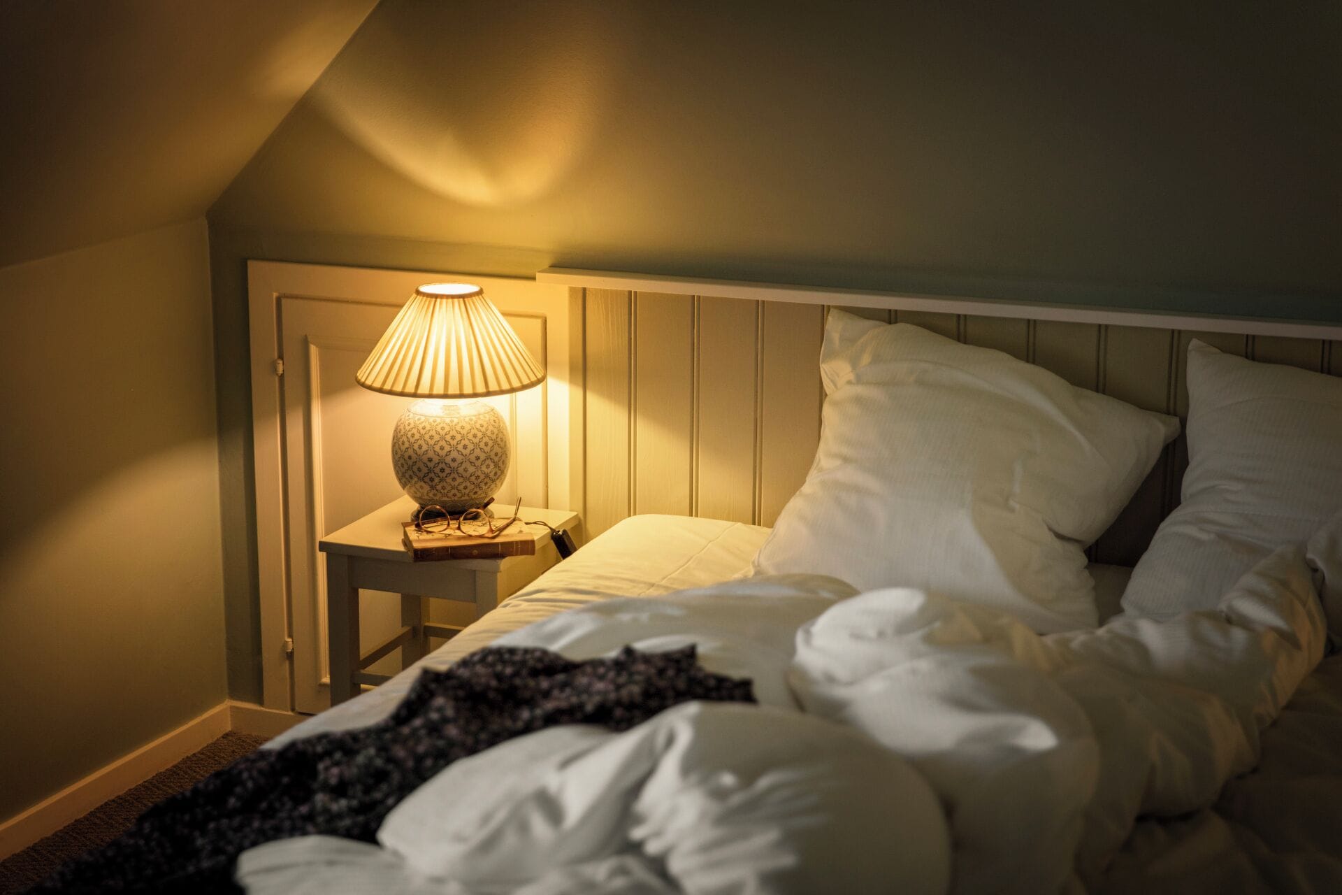 Ein Schlafzimmer mit einem Bett, weißen Kissen, einem Nachttisch mit Lampe und einem Buch.