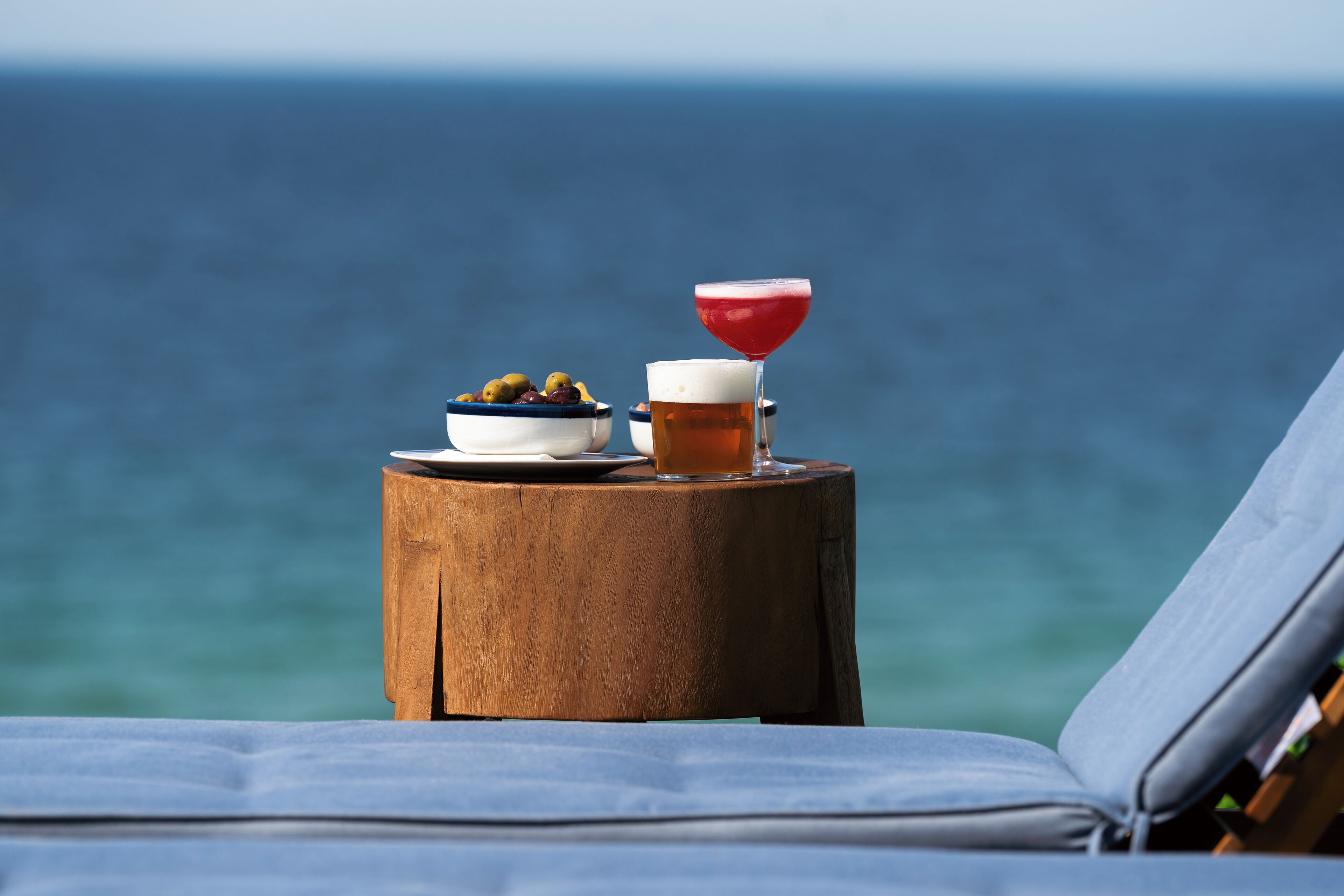 A wooden table with a bowl of olives and a glass of beer, set on a beach with a lounge chair and ocean view.