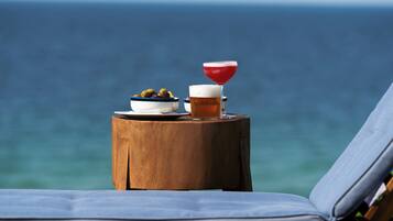 Ein Holztisch mit einer Oliven-Schale und einem Bierglas, platziert an einem Strand mit Liegestuhl und Meerblick.