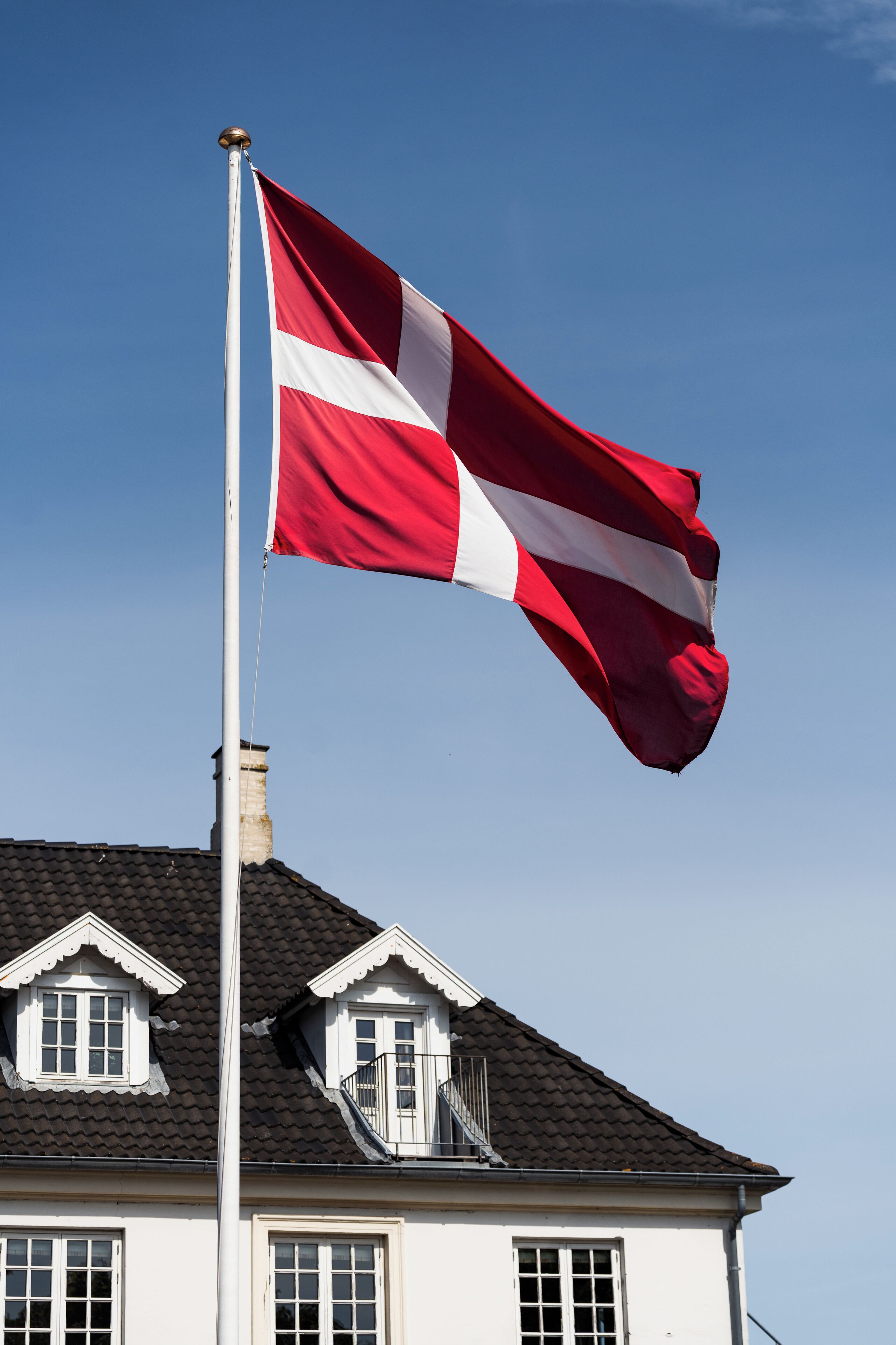Et flagstang med flag, et hus med mørkt tag og en klar blå himmel.
