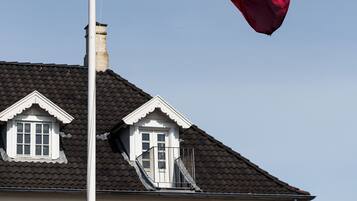 Et flagstang med flag, et hus med mørkt tag og en klar blå himmel.