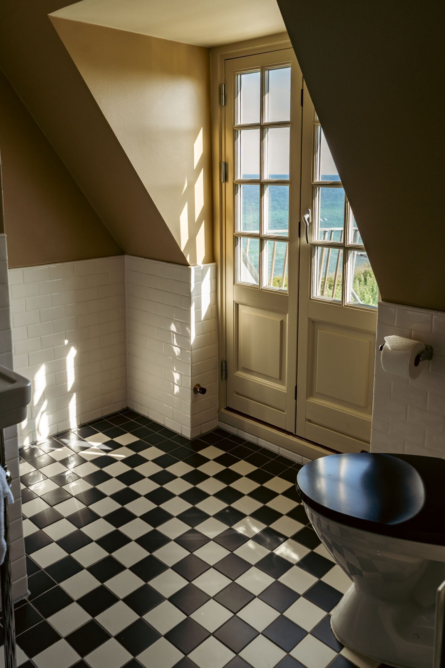 Een badkamer met een zwart-wit geruite vloer, witte betegelde wanden, een witte deur met glaspanelen en uitzicht op zee door de deur.