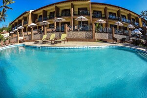 Un hotel con zona de piscina, múltiples sillas de descanso, sombrillas y un edificio de dos plantas con balcones.