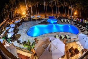 A nighttime swimming pool area with lounge chairs and umbrellas, surrounded by palm trees and a beach in the background.