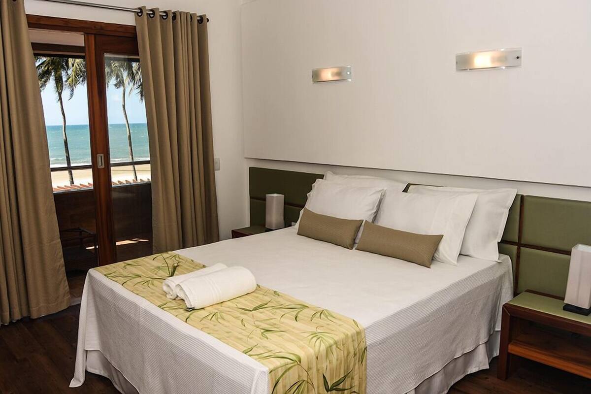 A hotel room with a bed, a nightstand, a lamp, and a view of the beach through a sliding door.