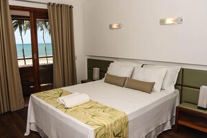 Habitación de hotel con cama, mesita de noche, lámpara y vista a la playa a través de una puerta corrediza.