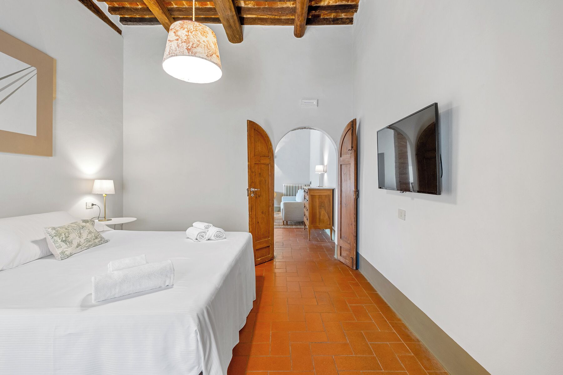 Un dormitorio con una cama, una mesita de noche con lámpara, una toalla enrollada, una puerta de madera y un televisor montado en la pared.