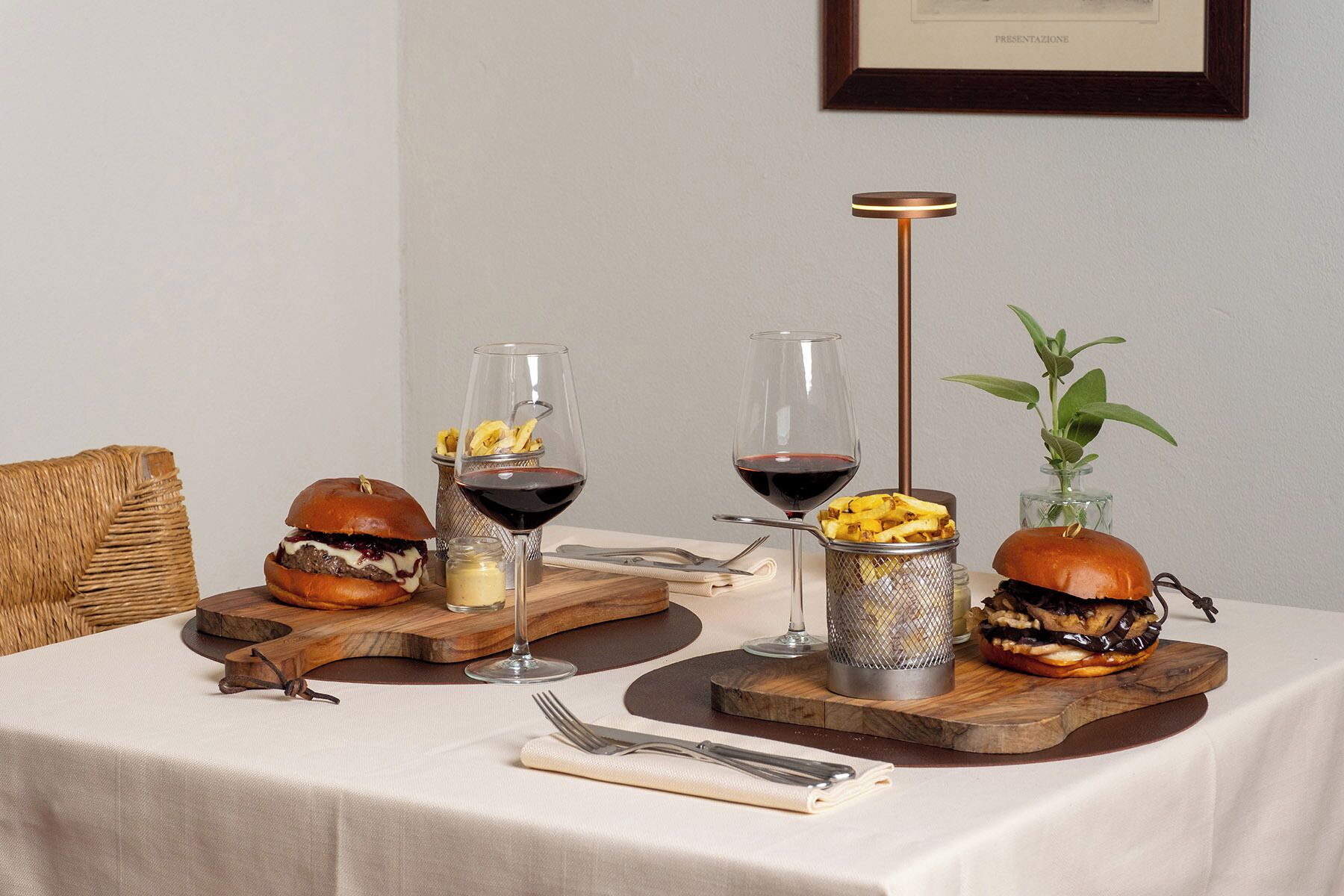 A dining table with two burgers, two glasses of red wine, a jar of sauce, and a plant.