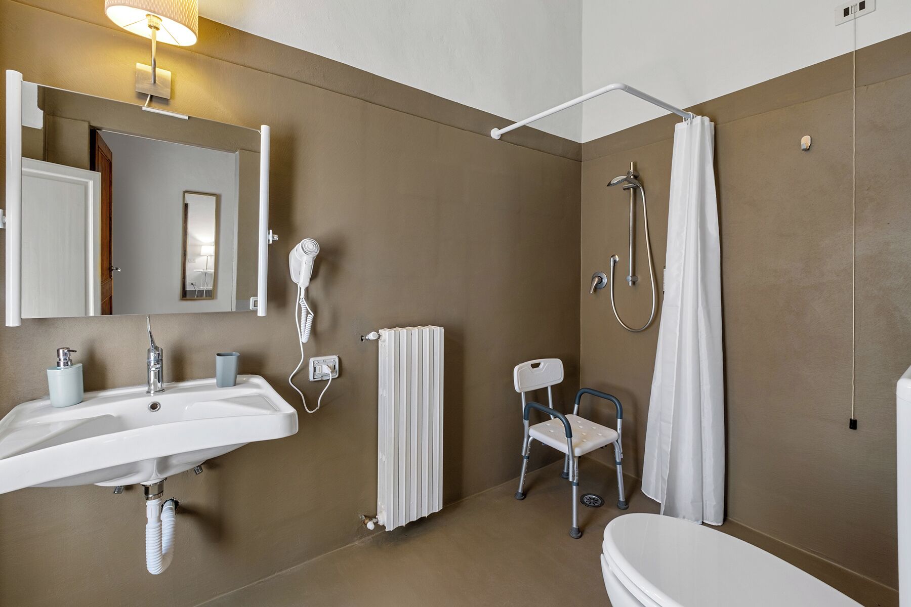 A bathroom with a sink, mirror, shower, and a wheelchair-accessible chair.