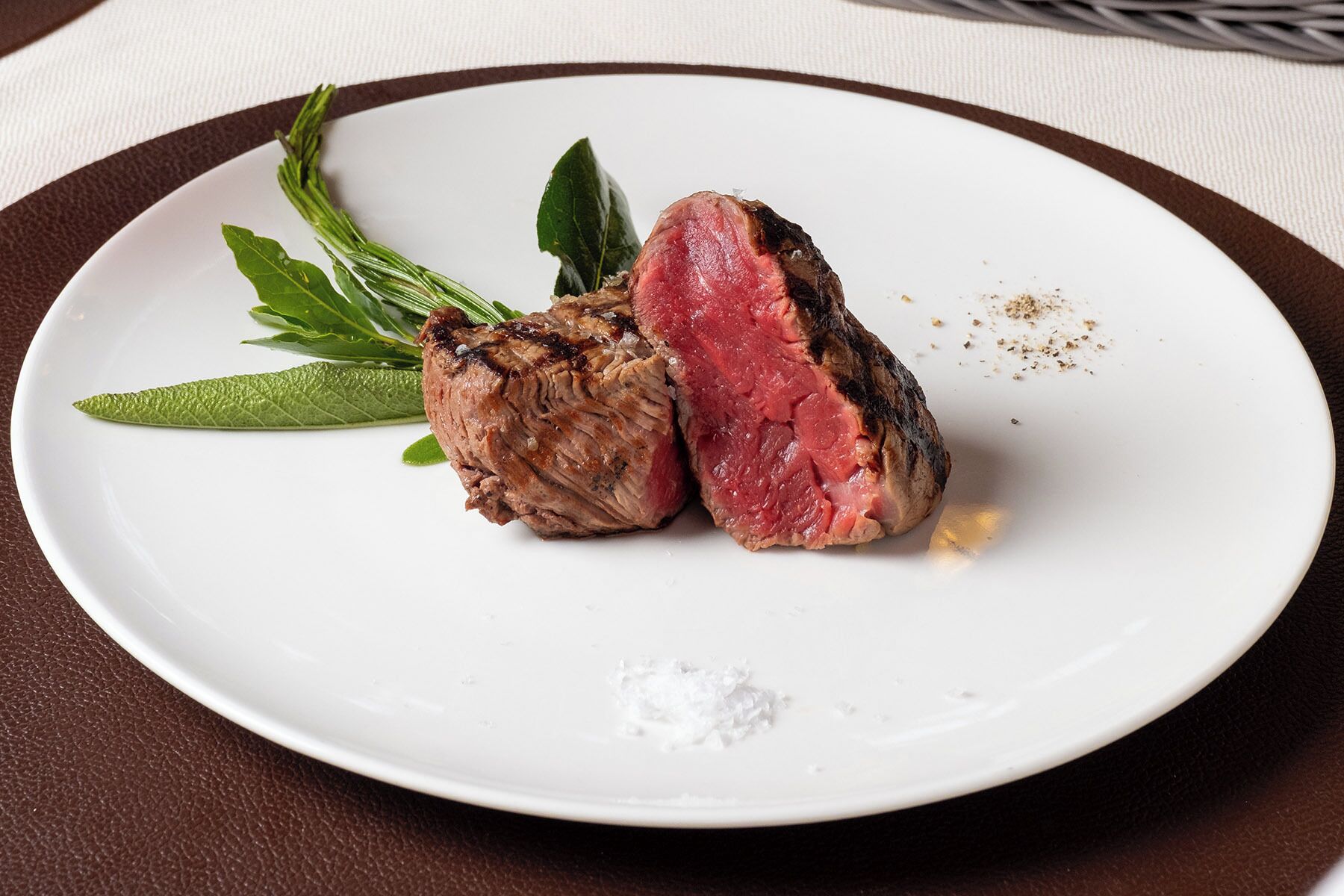 A white plate with a grilled steak, a sprig of rosemary, and a small pile of salt.