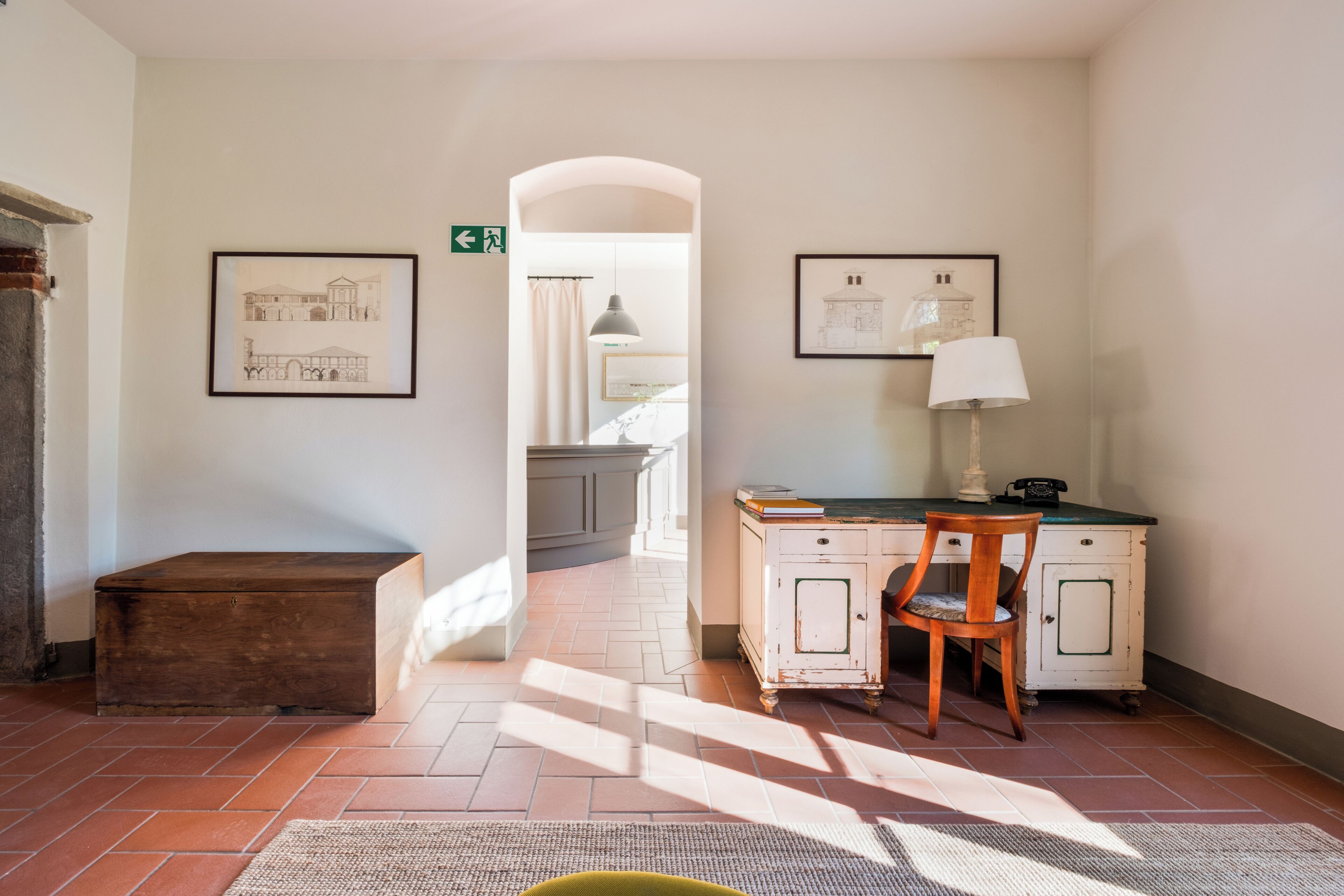 A hallway with a wooden bench, a desk with a lamp, and framed architectural drawings on the walls.