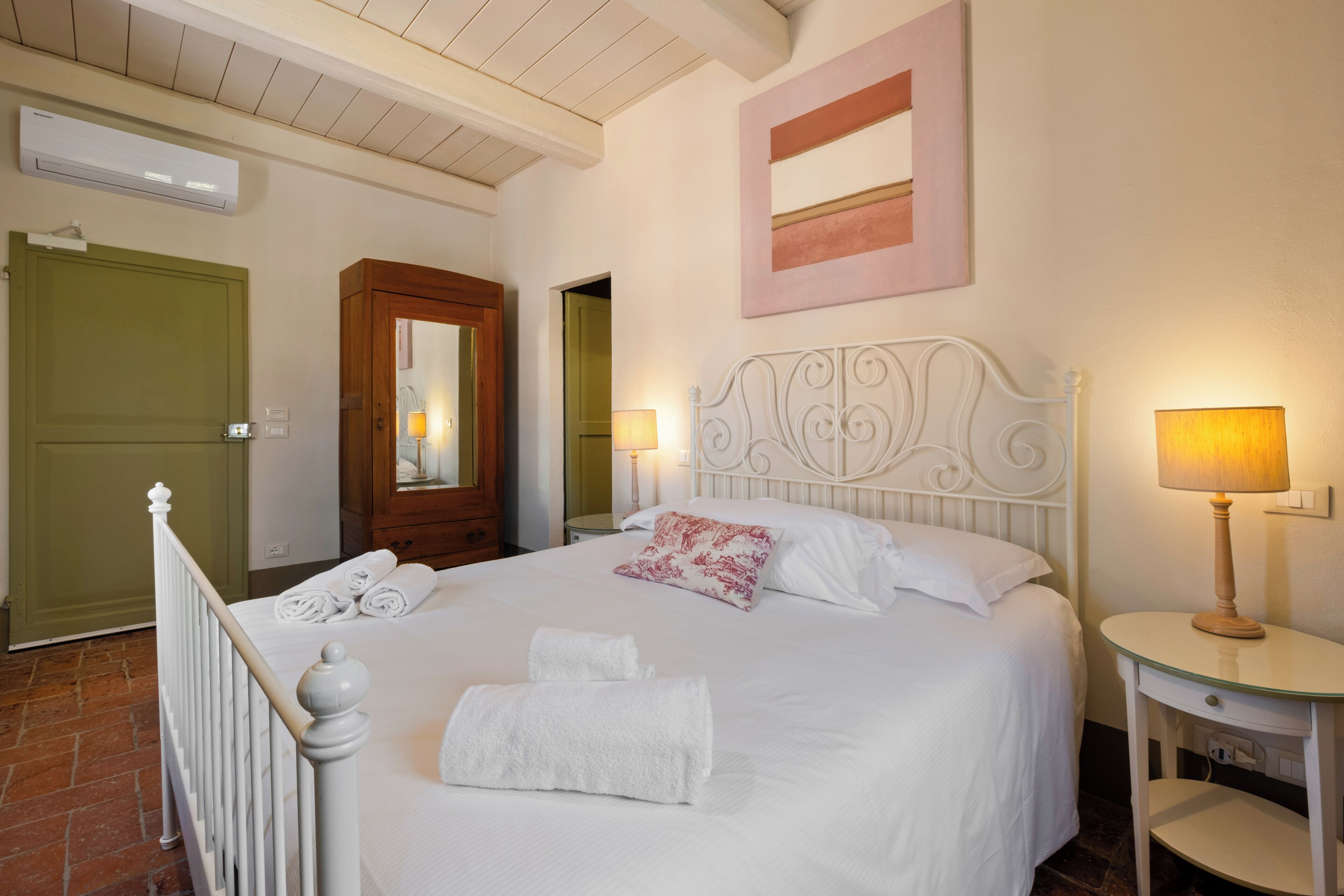 A neatly made bed with white linens and two rolled towels, a bedside lamp, and a wooden door.