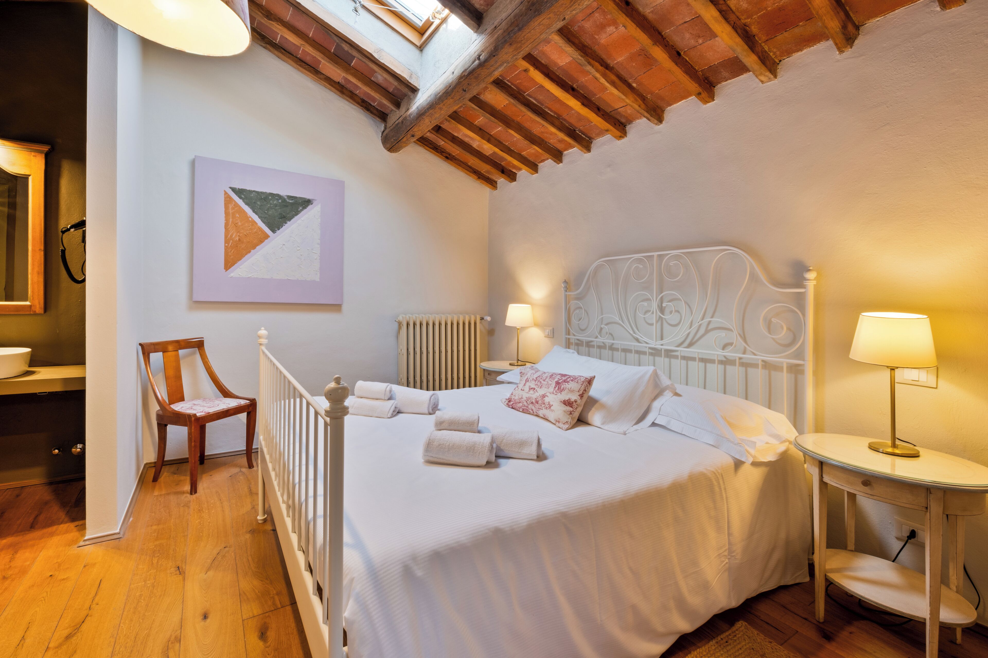 A bedroom with a bed, two bedside tables, a chair, a radiator, and a painting on the wall.