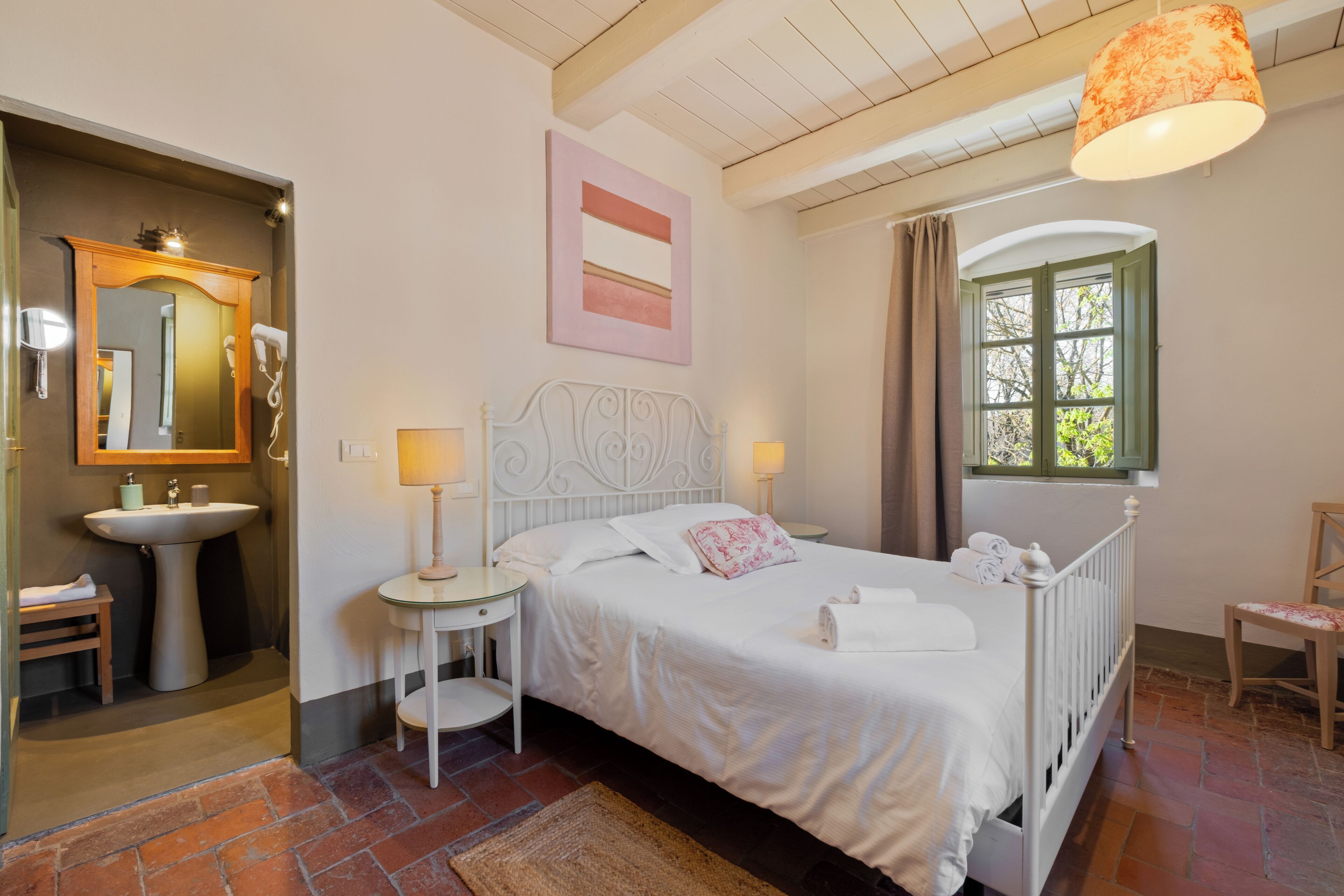 Un dormitorio con cama, mesitas de noche, espejo, ventana y baño con lavabo.