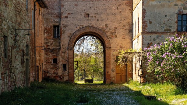 An arched stone gateway between two buildings, a grassy area, and a flowering bush. - Tenuta la Fratta (Sinalunga)