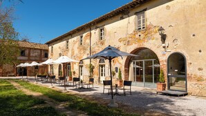 An outdoor seating area with tables and chairs, a gravel path, and a building with arched doorways. - Tenuta la Fratta (Sinalunga)